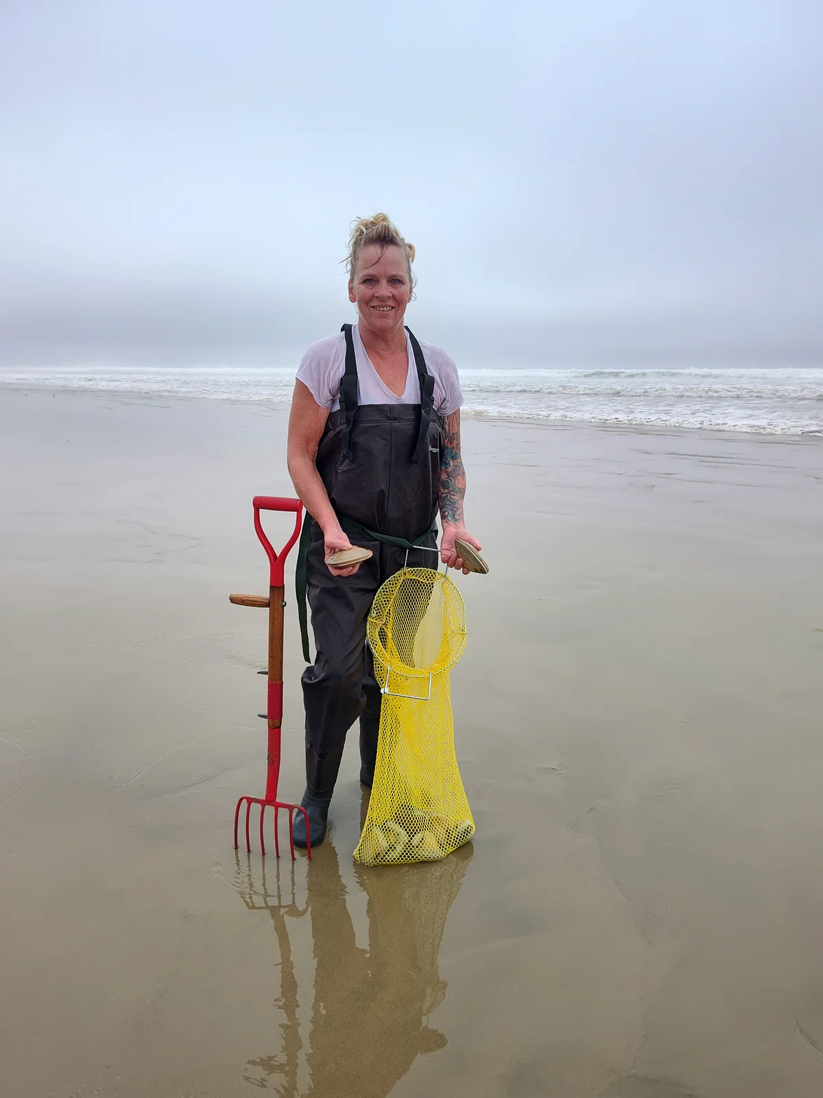 A clammer in chest waders standing on the wet sand at Pismo Beach on a foggy morning, holding two large clams above a yellow mesh catch bag, with a red clam fork planted upright beside her.